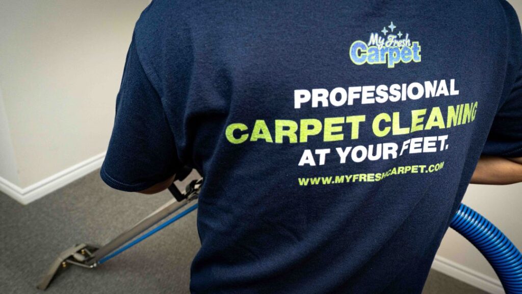 My Fresh Carpet worker cleaning the carpet with the wand tool while having his back facing the camera. His shirt says Professional Carpet Cleaning At Your Feet.